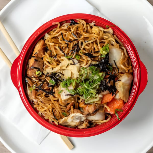 Yakisoba noodles with chicken, vegetables, and seaweed in a red bowl with chopsticks.