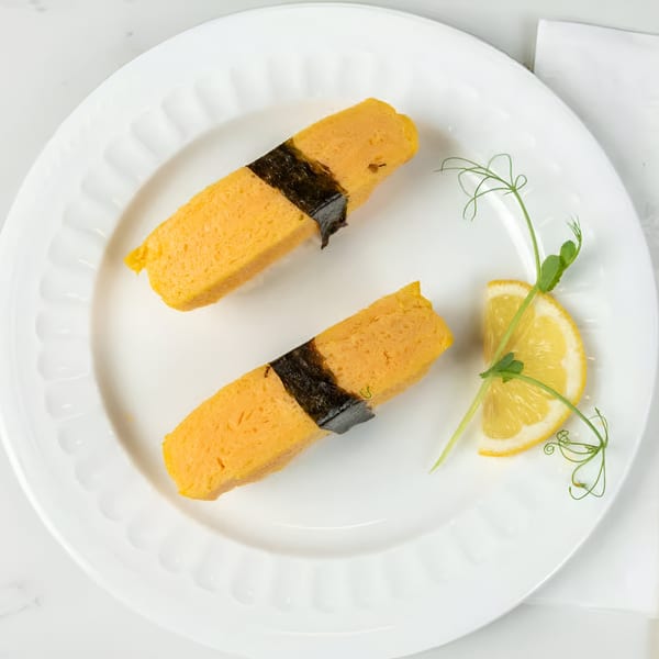 Two pieces of tamago nigiri, wrapped in nori, with a lemon wedge and microgreens on a white plate.